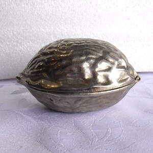 May include: A silver-colored metal nutcracker shaped like a walnut. The nutcracker is closed and has a detailed, textured surface.
