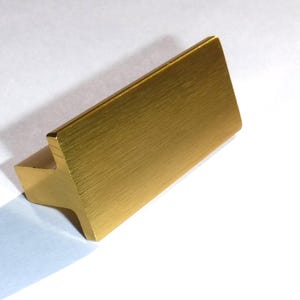 May include: A gold-colored, rectangular cabinet pull with a brushed finish. The pull has a T-shaped design, with a flat, horizontal bar and a vertical extension. The hardware is made of metal and is suitable for drawers or cabinets.