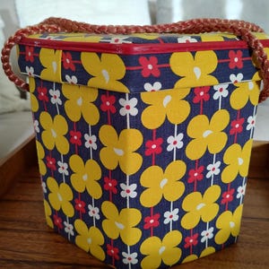 May include: A vintage-style box with a navy blue background and a repeating pattern of large yellow flowers, red and white smaller flowers. The box has a red trim and a braided brown handle.