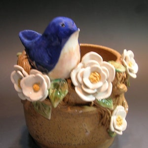 May include: A ceramic planter with a blue bird perched on the rim, surrounded by white flowers and green leaves. The planter is brown with a speckled glaze.