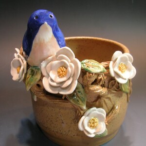 May include: A ceramic planter with a blue bird perched on the rim, surrounded by white flowers and green leaves. The planter is brown with a speckled glaze.