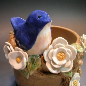 May include: A ceramic bird figurine with blue and pink painted details, perched on a brown ceramic base with white ceramic flowers.