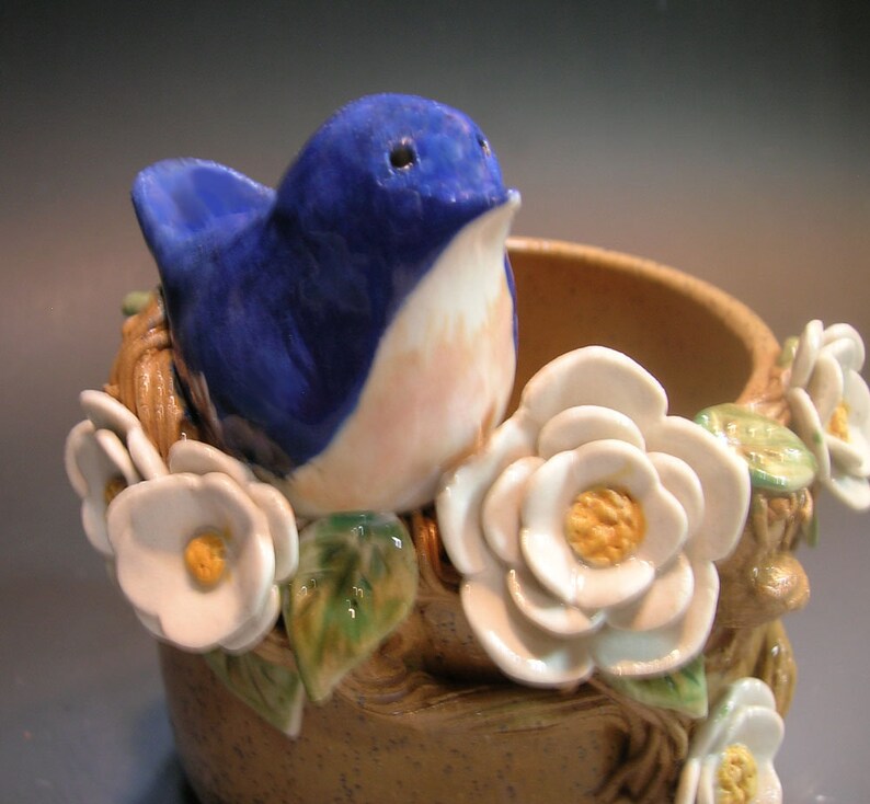 May include: A ceramic bird figurine with blue and pink painted details, perched on a brown ceramic base with white ceramic flowers.