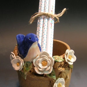 May include: A ceramic pencil holder shaped like a bird's nest with a blue bird, white flowers, and green leaves. The holder has a brown speckled glaze and is filled with pencils with white polka dots.