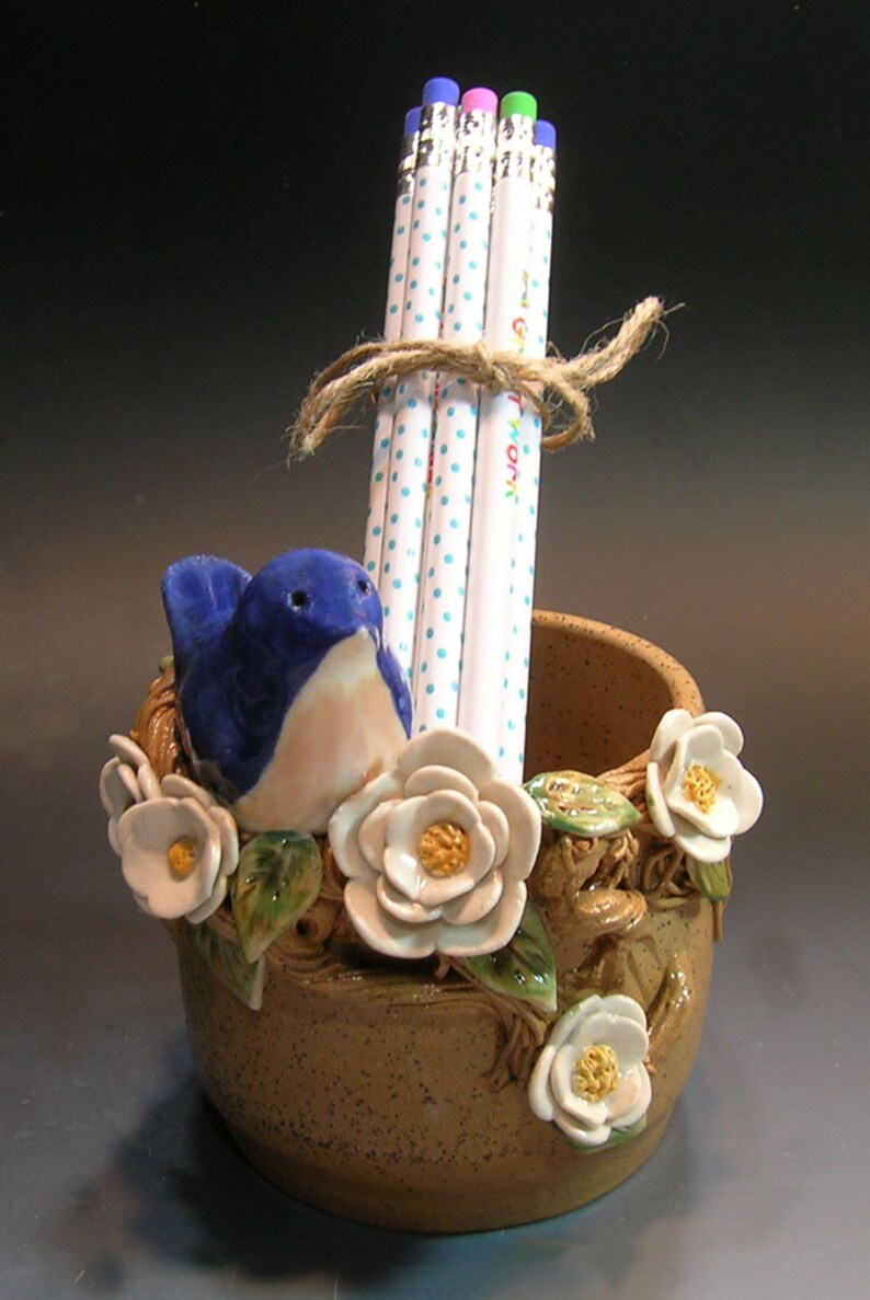 May include: A ceramic pencil holder shaped like a bird's nest with a blue bird, white flowers, and green leaves. The holder has a brown speckled glaze and is filled with pencils with white polka dots.