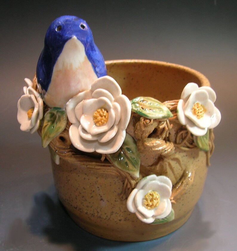 May include: A ceramic planter with a blue bird perched on the rim, surrounded by white flowers and green leaves. The planter is brown with a speckled glaze.