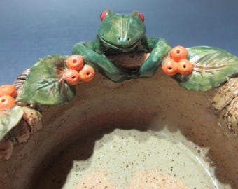 May include: A ceramic bowl with a green frog and orange berries on the rim. The bowl is made to look like a woven basket.