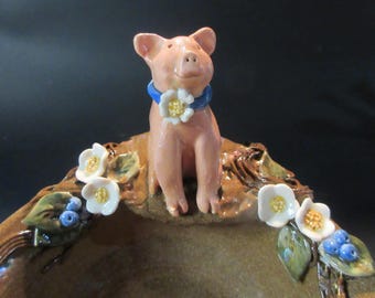 May include: A ceramic pig figurine with a blue ribbon and white flowers around its neck, sitting on a brown ceramic dish with white flowers and blue berries.
