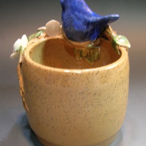May include: A ceramic planter with a blue bird perched on the rim. The planter is speckled brown and has a small opening at the top. The bird is perched on a green leafy branch.