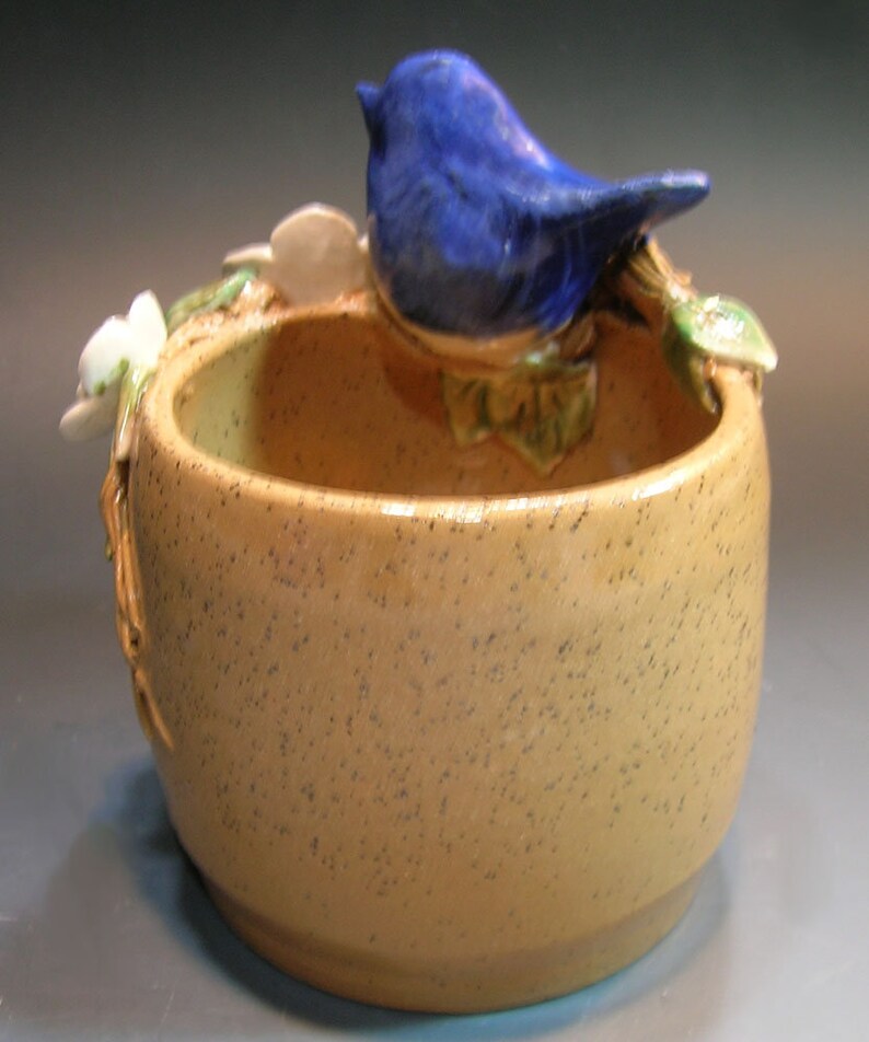 May include: A ceramic planter with a blue bird perched on the rim. The planter is speckled brown and has a small opening at the top. The bird is perched on a green leafy branch.