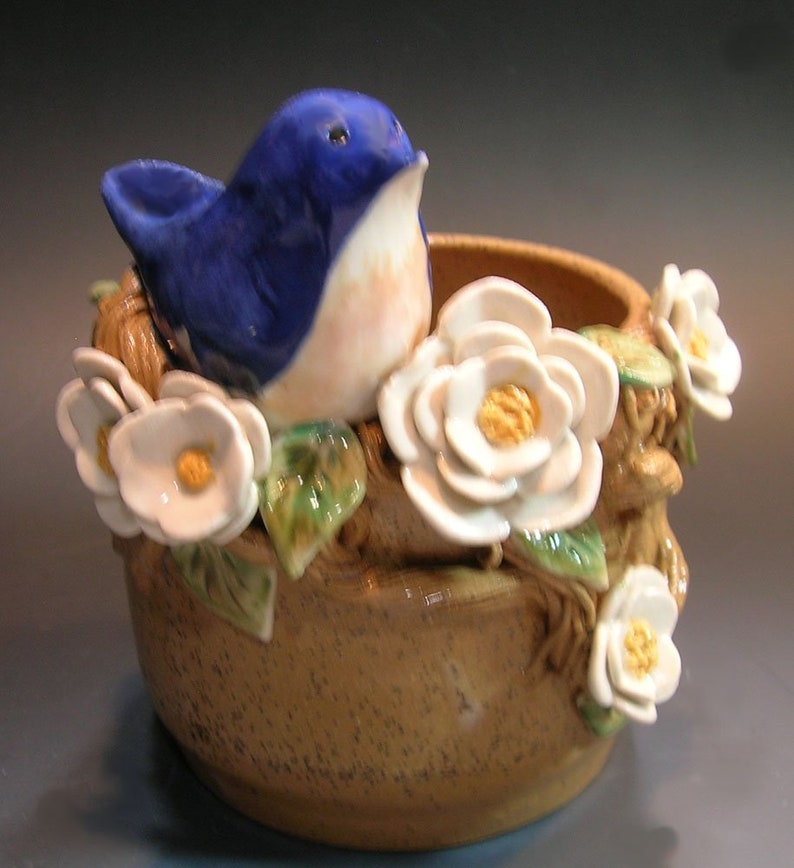 May include: A ceramic planter with a blue bird perched on the rim, surrounded by white flowers and green leaves. The planter is brown with a speckled glaze.
