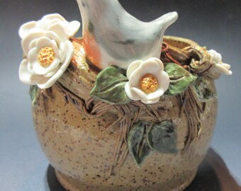 May include: A ceramic bird perched on a brown and white speckled pot with white flowers and green leaves. The bird is light blue with a white belly and a small hole on its head.