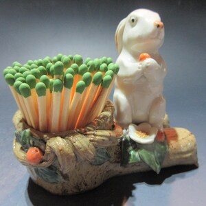 May include: A ceramic match holder shaped like a log with a white rabbit figurine sitting on top. The rabbit is holding a small orange carrot and is surrounded by green leaves. The match holder has a brown and green glaze with orange accents.