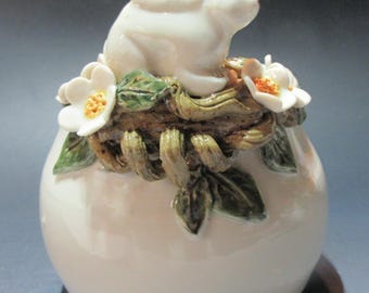 May include: A white ceramic vase with a white rabbit sitting on top of it. The vase is decorated with green leaves and white flowers.