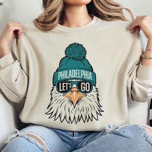 May include: A beige sweatshirt featuring a graphic of an eagle wearing a teal beanie with the words "Philadelphia Let's Go" on it. The eagle is also wearing sunglasses.