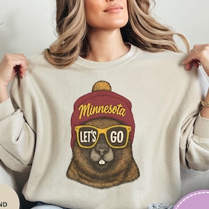 May include: Sand-colored sweatshirt featuring a graphic of a gopher wearing a maroon beanie and yellow sunglasses. The text "Minnesota LET'S GO" is arched above the gopher. The sweatshirt is a casual style.