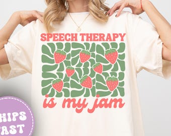 May include: Ivory-colored t-shirt with the text "SPEECH THERAPY is my jam" in pink and a green and pink strawberry design. The shirt is a relaxed fit.