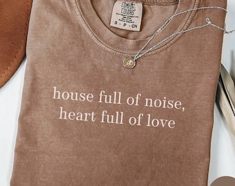 May include: A brown t-shirt with the text "house full of noise, heart full of love" printed in white. The shirt is made of a soft, comfortable material and has a relaxed fit. The shirt is shown on a white background with a hat and a necklace.