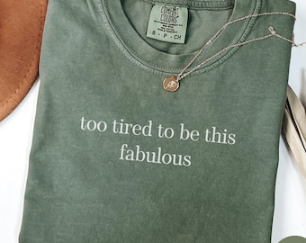 May include: A moss green t-shirt with the phrase "too tired to be this fabulous" in white lettering. A gold necklace with a pendant rests on the shirt. The shirt is folded and displayed on a white surface.
