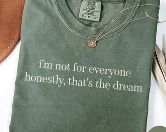 May include: A moss green t-shirt with the text "i'm not for everyone honestly, that's the dream" in white. A gold necklace with a pendant is draped over the shirt. The shirt has a Comfort Colors tag.