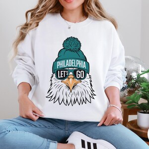 May include: White sweatshirt featuring a cartoon eagle wearing a teal beanie with the text "Philadelphia Let's Go" and sunglasses.