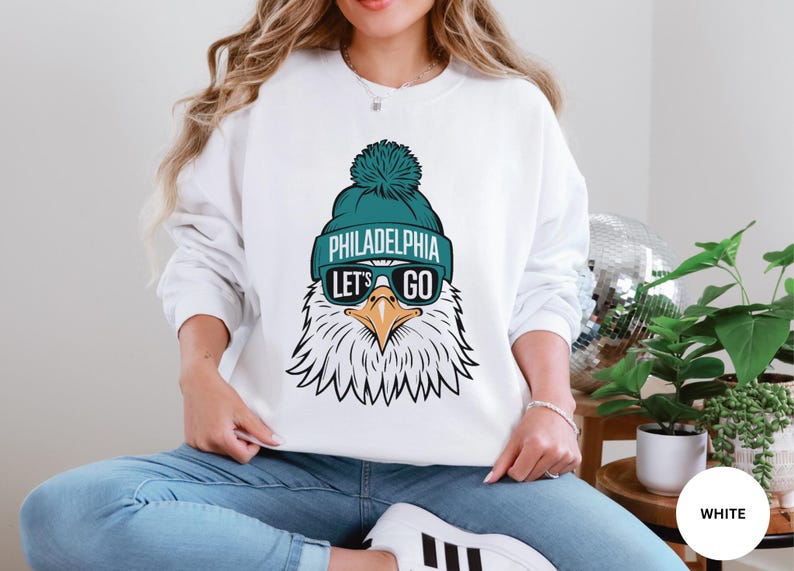 May include: White sweatshirt featuring a cartoon eagle wearing a teal beanie with the text "Philadelphia Let's Go" and sunglasses.