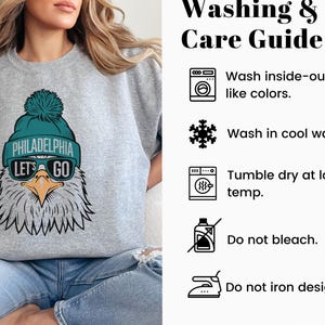 May include: Grey sweatshirt featuring a teal and white eagle graphic. The eagle is wearing a teal hat with the words "Philadelphia" and "Let's Go" on it. The sweatshirt includes a care guide with instructions for washing, drying, and ironing.