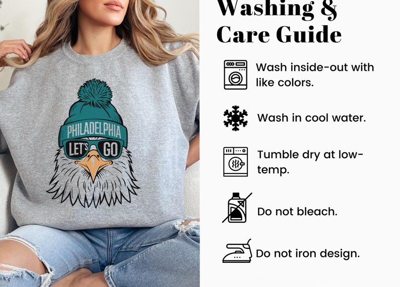 May include: Grey sweatshirt featuring a teal and white eagle graphic. The eagle is wearing a teal hat with the words "Philadelphia" and "Let's Go" on it. The sweatshirt includes a care guide with instructions for washing, drying, and ironing.