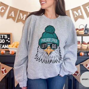 May include: A grey sweatshirt featuring a graphic of a Philadelphia Eagles mascot wearing a green and white hat and sunglasses. The text "Philadelphia" and "Let's Go" is printed on the sweatshirt.
