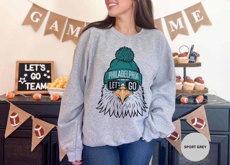 May include: A grey sweatshirt featuring a graphic of a Philadelphia Eagles mascot wearing a green and white hat and sunglasses. The text "Philadelphia" and "Let's Go" is printed on the sweatshirt.