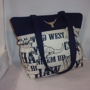 Puede incluir: Una bolsa de mano azul marino y blanca con tem&aacute;tica western. La bolsa presenta una cabeza de toro longhorn, las palabras "Wild West", "Cowboy", "Haw", y "Ride 'em Up".