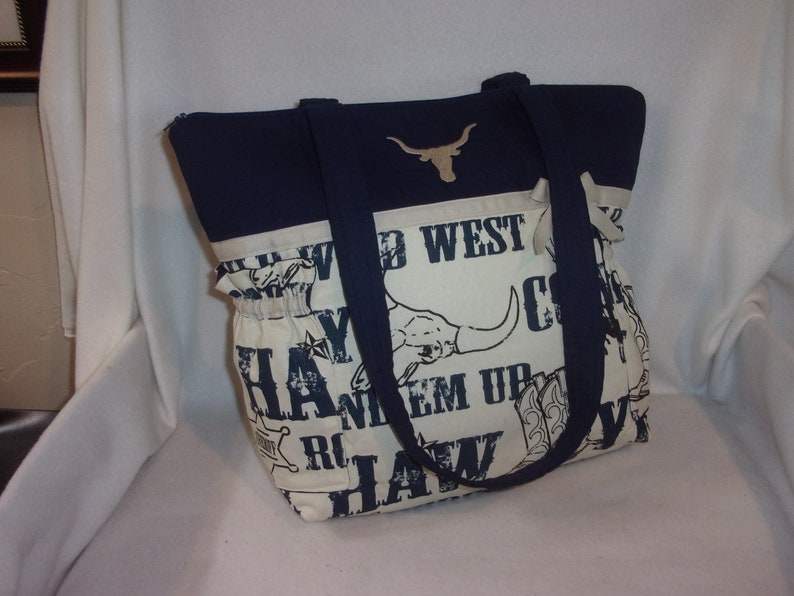 Puede incluir: Una bolsa de mano azul marino y blanca con tem&aacute;tica western. La bolsa presenta una cabeza de toro longhorn, las palabras "Wild West", "Cowboy", "Haw", y "Ride 'em Up".