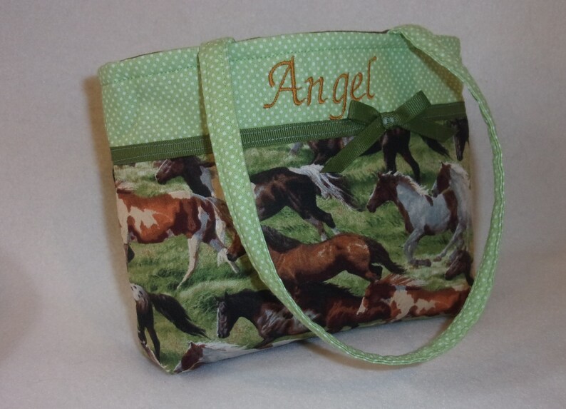 Pode incluir: Pequena sacola com tecido tem&aacute;tico de cavalos. A sacola apresenta acabamento e al&ccedil;as de bolinhas verdes, com o nome "Angel" bordado em fio dourado. Um la&ccedil;o verde adiciona um toque decorativo.