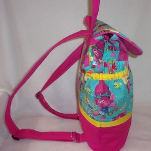 May include: A pink backpack with adjustable straps. The front of the backpack features a turquoise fabric with a colourful print of cartoon trolls. The print includes the words "Trolls" and "Just Be You".