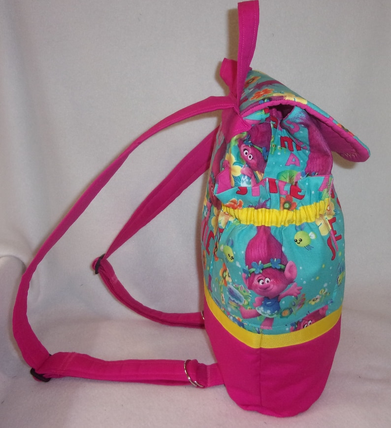 May include: A pink backpack with adjustable straps. The front of the backpack features a turquoise fabric with a colourful print of cartoon trolls. The print includes the words "Trolls" and "Just Be You".