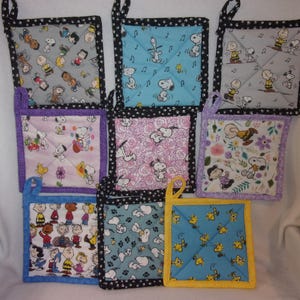 May include: A collection of square quilted pot holders featuring various Peanuts characters. Each pot holder has a different patterned fabric, including Snoopy, Charlie Brown, and Woodstock, with coordinating trim and a hanging loop. The pot holders are in various colors.
