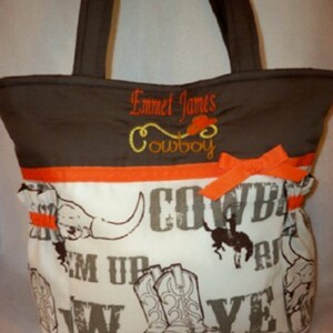 Puede incluir: Una bolsa de mano marr&oacute;n y blanca con tem&aacute;tica vaquera. La bolsa tiene una solapa marr&oacute;n con el nombre "Emmet James Cowboy" bordado. La parte principal de la bolsa es blanca con un estampado vaquero marr&oacute;n y naranja.