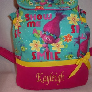 May include: A pink and turquoise backpack with a yellow ribbon. The backpack features a cartoon troll with pink hair and the text "Show Me A Smile". The backpack is personalised with the name "Kayleigh" in yellow embroidery.