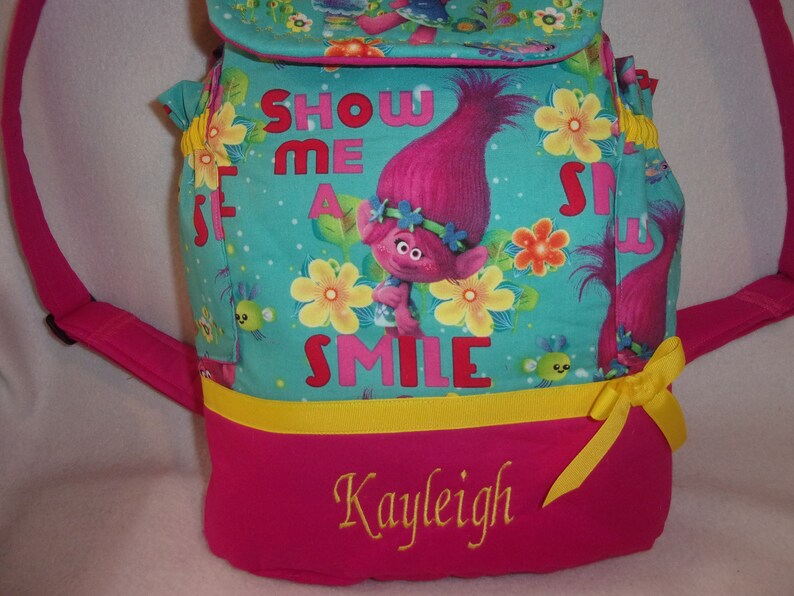 May include: A pink and turquoise backpack with a yellow ribbon. The backpack features a cartoon troll with pink hair and the text "Show Me A Smile". The backpack is personalised with the name "Kayleigh" in yellow embroidery.