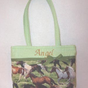Pode incluir: Uma sacola com a parte superior e al&ccedil;as de bolinhas verdes. O corpo principal da sacola apresenta uma estampa de cavalos correndo em v&aacute;rias cores. O nome "Angel" &eacute; bordado em uma fonte cursiva.