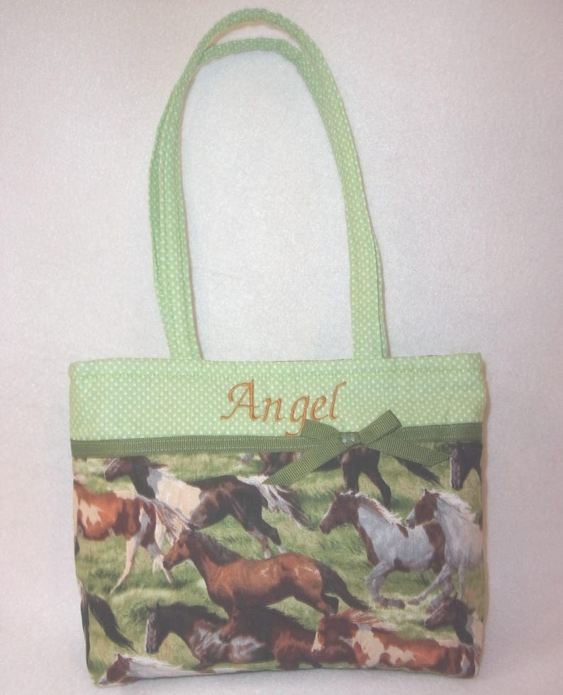 Pode incluir: Uma sacola com a parte superior e al&ccedil;as de bolinhas verdes. O corpo principal da sacola apresenta uma estampa de cavalos correndo em v&aacute;rias cores. O nome "Angel" &eacute; bordado em uma fonte cursiva.