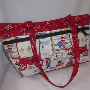 May include: Red and white fabric tote bag with a western theme. The bag features a red and white bandana print, black trim, and a scene of cowboys and a wagon.