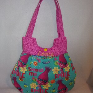 May include: Pink and teal fabric tote bag with a cartoon troll character and the words "Show Me A Smile". The bag has a pink fabric lining and the name "Leena" embroidered on the front.