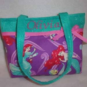 May include: Purple tote bag with a pink ribbon and teal straps. The bag features a print of Ariel from The Little Mermaid. The bag is personalised with the name "Olivia" in teal embroidery.