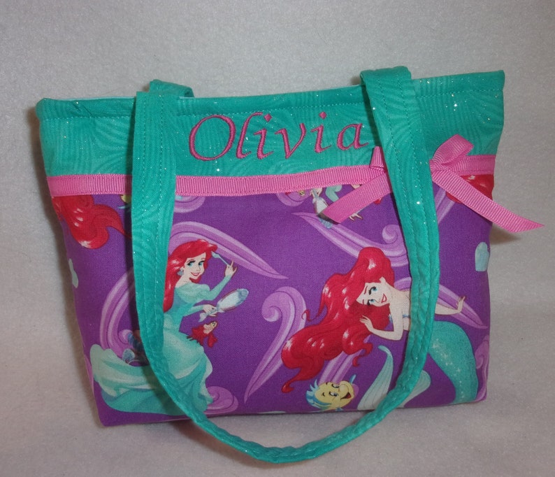 May include: Purple tote bag with a pink ribbon and teal straps. The bag features a print of Ariel from The Little Mermaid. The bag is personalised with the name "Olivia" in teal embroidery.