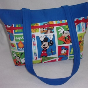 May include: A blue tote bag featuring a colorful cartoon print with Mickey Mouse, Goofy, Donald Duck, and other characters. The bag has blue handles and trim, with the word "EXPLORE" printed on the front.