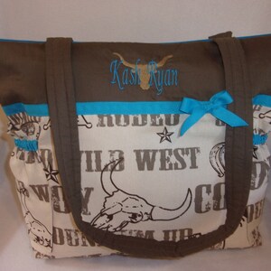 Puede incluir: Una bolsa de mano marr&oacute;n y turquesa con un panel de tela blanca con un tema occidental con las palabras "Wild West" y una calavera con cuernos. La bolsa tiene una cremallera marr&oacute;n y dos asas marrones. La bolsa tiene una cinta turquesa atada en un lazo en la parte delantera. La bolsa est&aacute; personalizada con el nombre "Kash Ryan" en bordado marr&oacute;n.