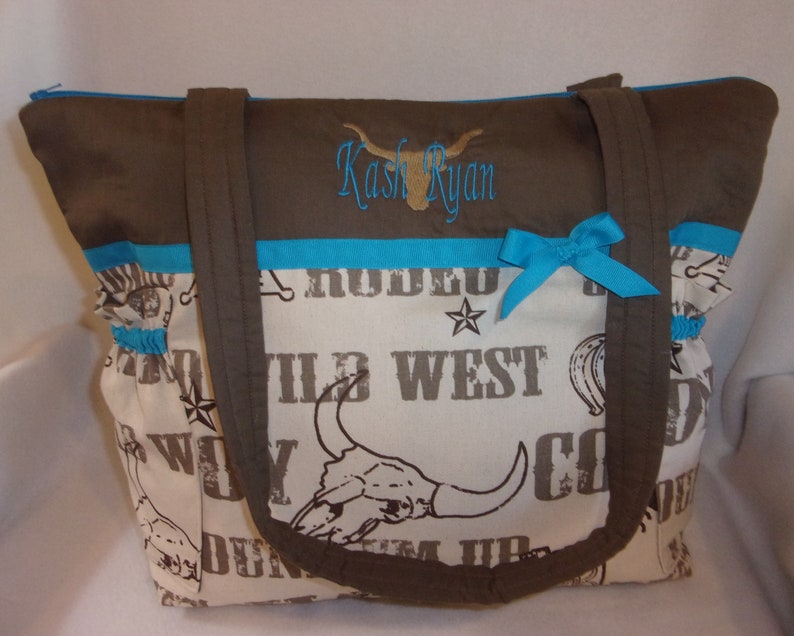 Puede incluir: Una bolsa de mano marr&oacute;n y turquesa con un panel de tela blanca con un tema occidental con las palabras "Wild West" y una calavera con cuernos. La bolsa tiene una cremallera marr&oacute;n y dos asas marrones. La bolsa tiene una cinta turquesa atada en un lazo en la parte delantera. La bolsa est&aacute; personalizada con el nombre "Kash Ryan" en bordado marr&oacute;n.