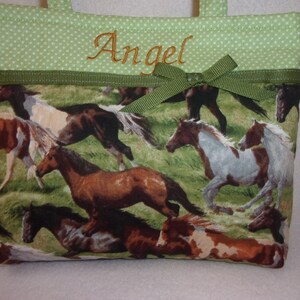 Pode incluir: Uma pequena sacola com um padr&atilde;o de cavalos a correr em tons de castanho, branco e bege sobre um fundo de relva verde. A parte superior &eacute; verde claro com bolinhas brancas e o nome "Angel" bordado em dourado.