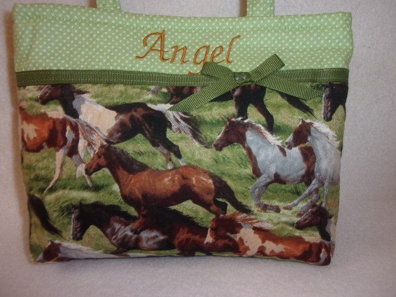 Pode incluir: Uma pequena sacola com um padr&atilde;o de cavalos a correr em tons de castanho, branco e bege sobre um fundo de relva verde. A parte superior &eacute; verde claro com bolinhas brancas e o nome "Angel" bordado em dourado.
